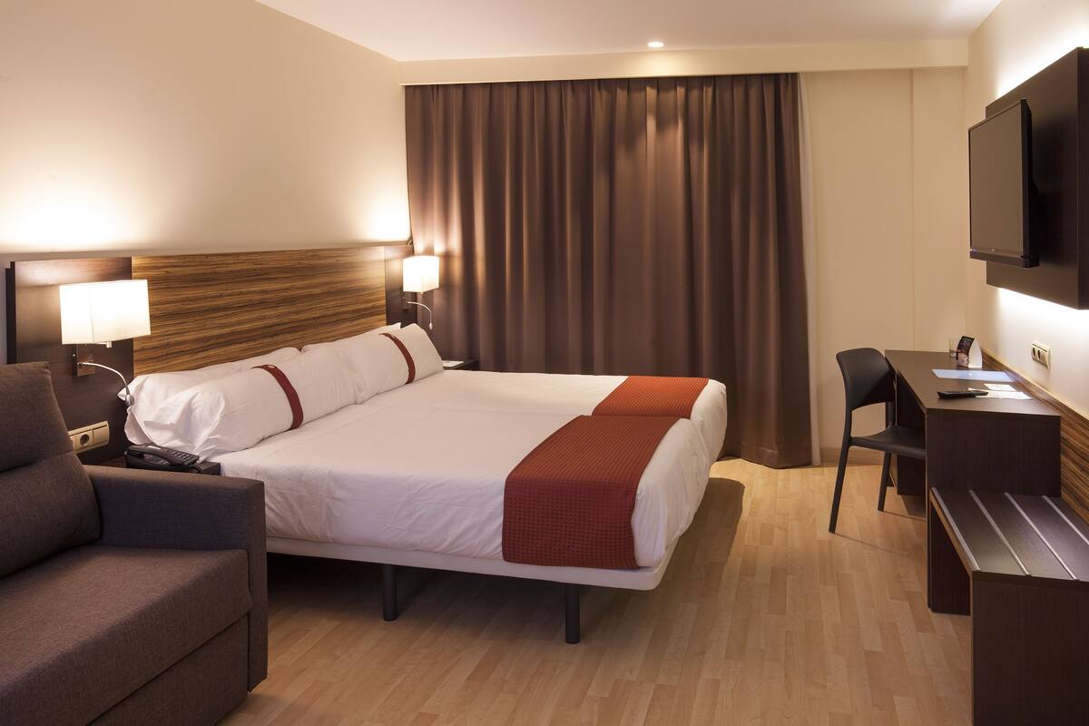 A hotel room with a large bed, a desk, a chair, a TV, and a lamp.