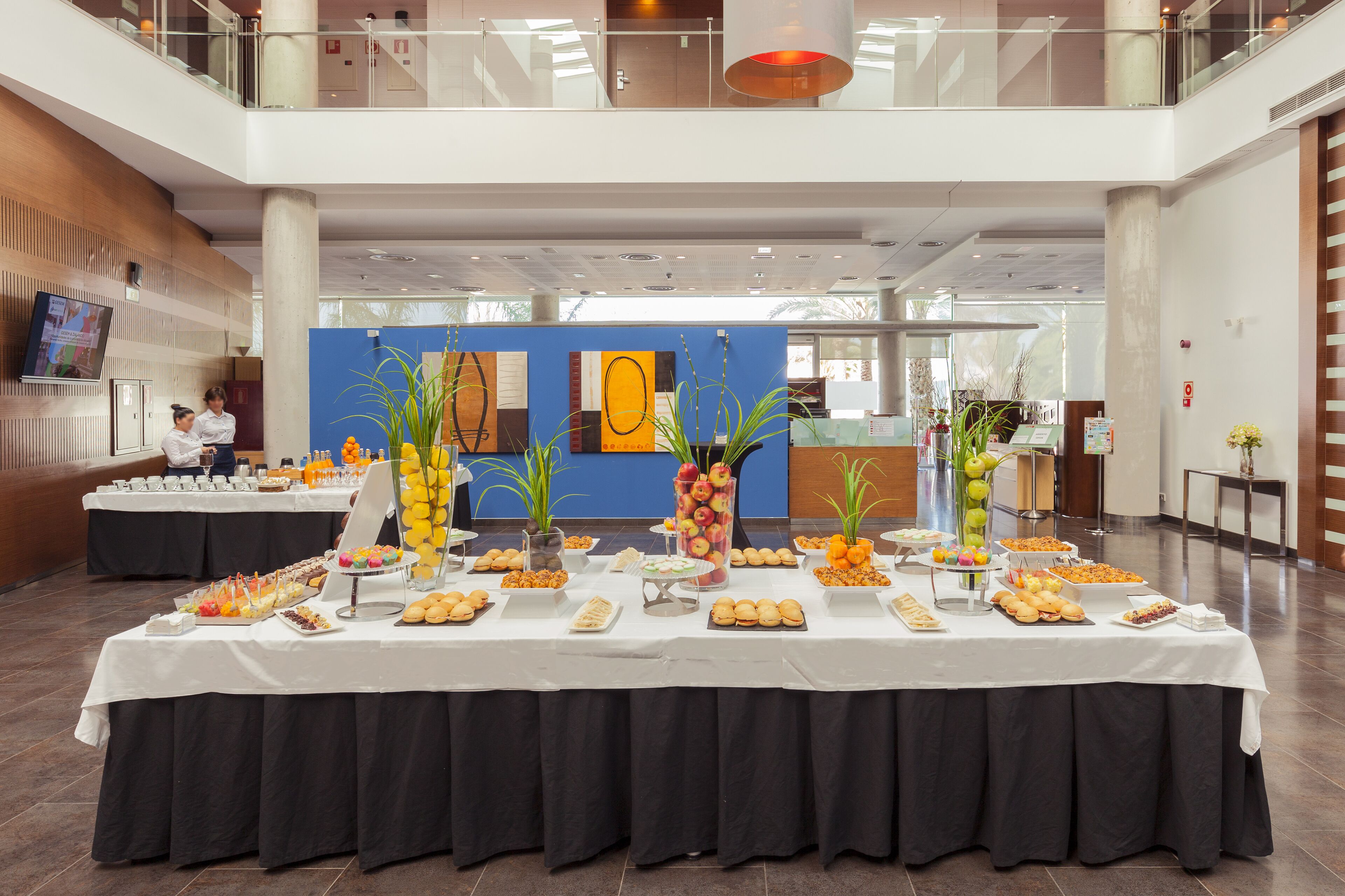 A buffet setup in a modern hotel lobby with multiple tables covered in food and drinks, and a large screen displaying content on the left.