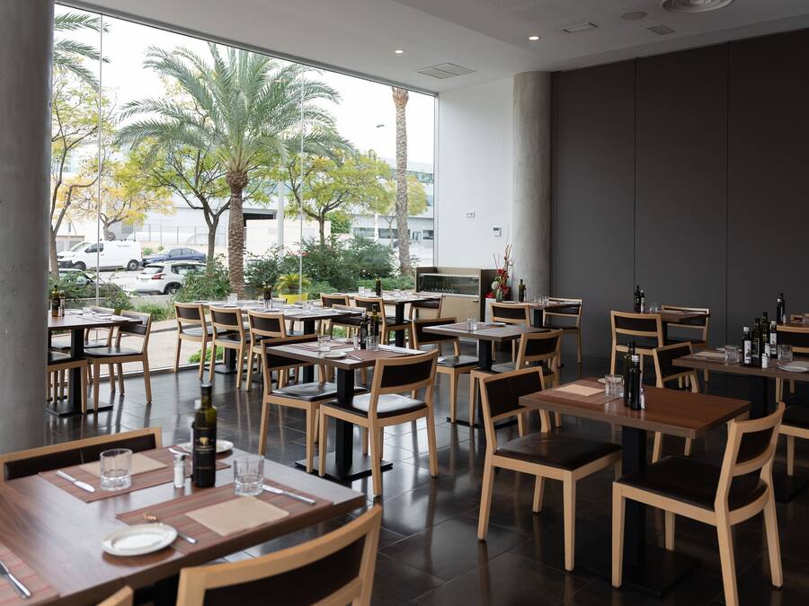 A restaurant with multiple tables and chairs, large windows offering a view of palm trees and parked cars outside.