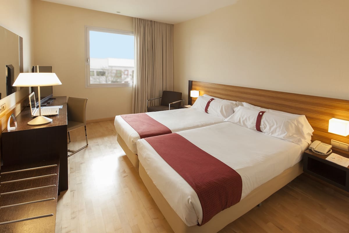 A hotel room with a double bed, a desk with a laptop, a chair, a nightstand with a phone, and a window with curtains.