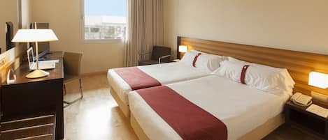 A hotel room with a double bed, a desk with a laptop, a chair, a nightstand with a phone, and a window with curtains.