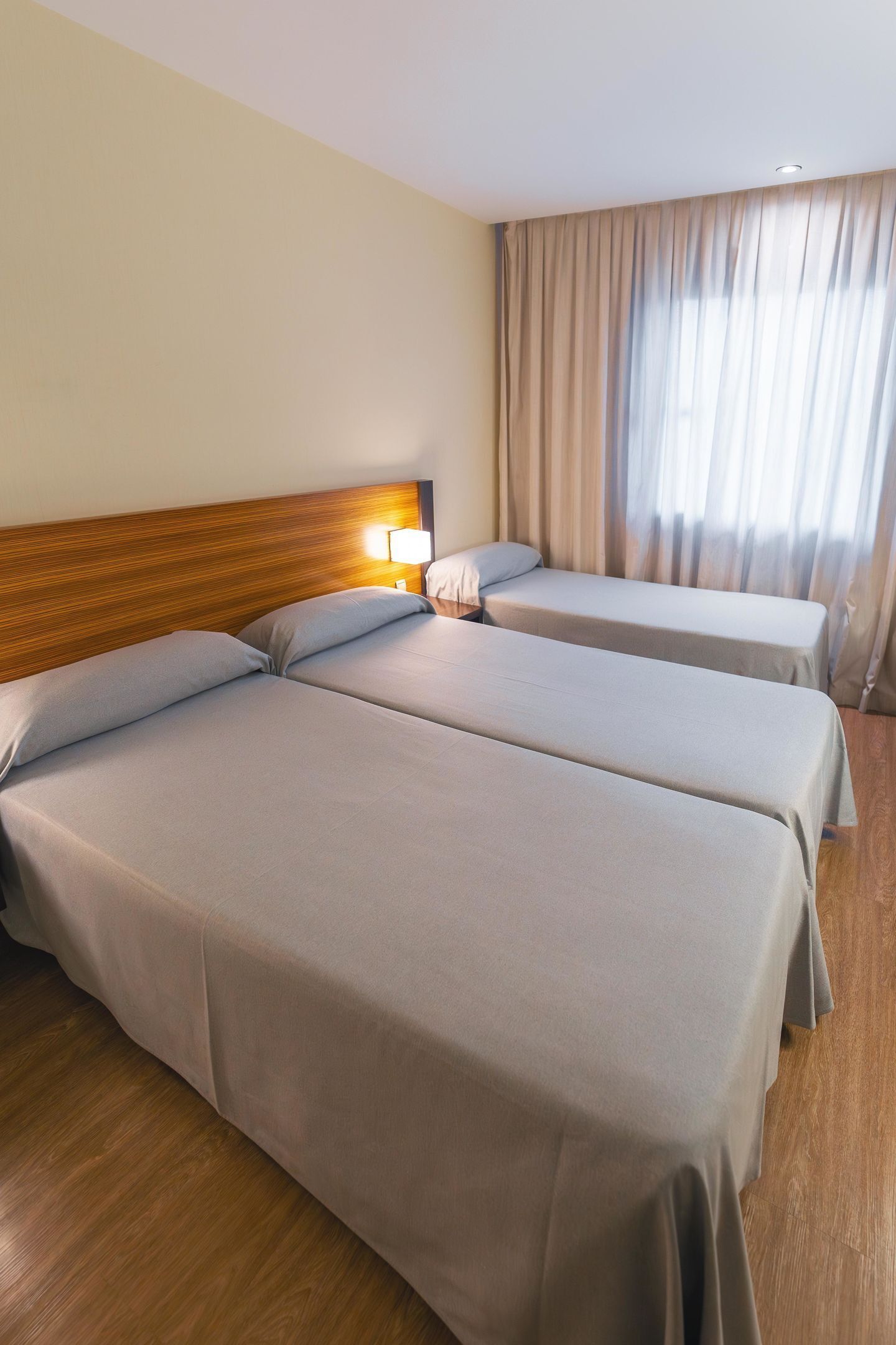 Habitación de hotel con dos camas individuales, cabeceros de madera, una mesita de noche con lámpara y un ventanal con cortinas.