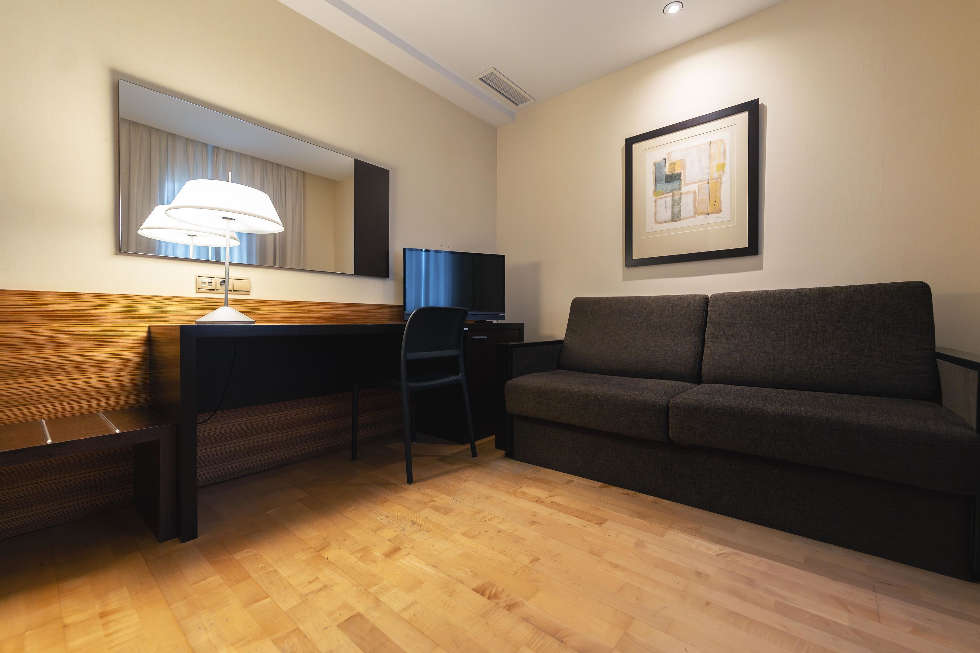 A hotel room with a wooden desk, a black chair, a grey sofa, a flat-screen TV, a wall-mounted lamp, and a framed picture on the wall.