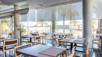 Un amplio espacio exterior con comedor, mesas, sillas, ventanas grandes y palmeras visibles.