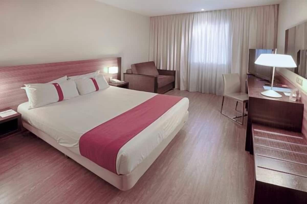 A hotel room with a large bed, a desk with a chair, a TV, and a window with curtains.