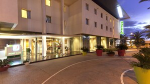 Un hotel con fachada moderna, una recepción visible y plantas en macetas alrededor de la entrada.