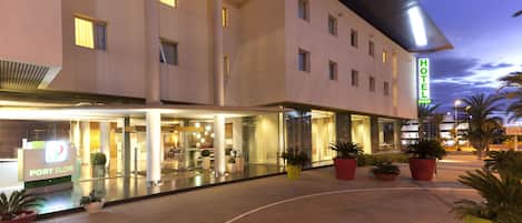 Un hotel con fachada moderna, una recepción visible y plantas en macetas alrededor de la entrada.