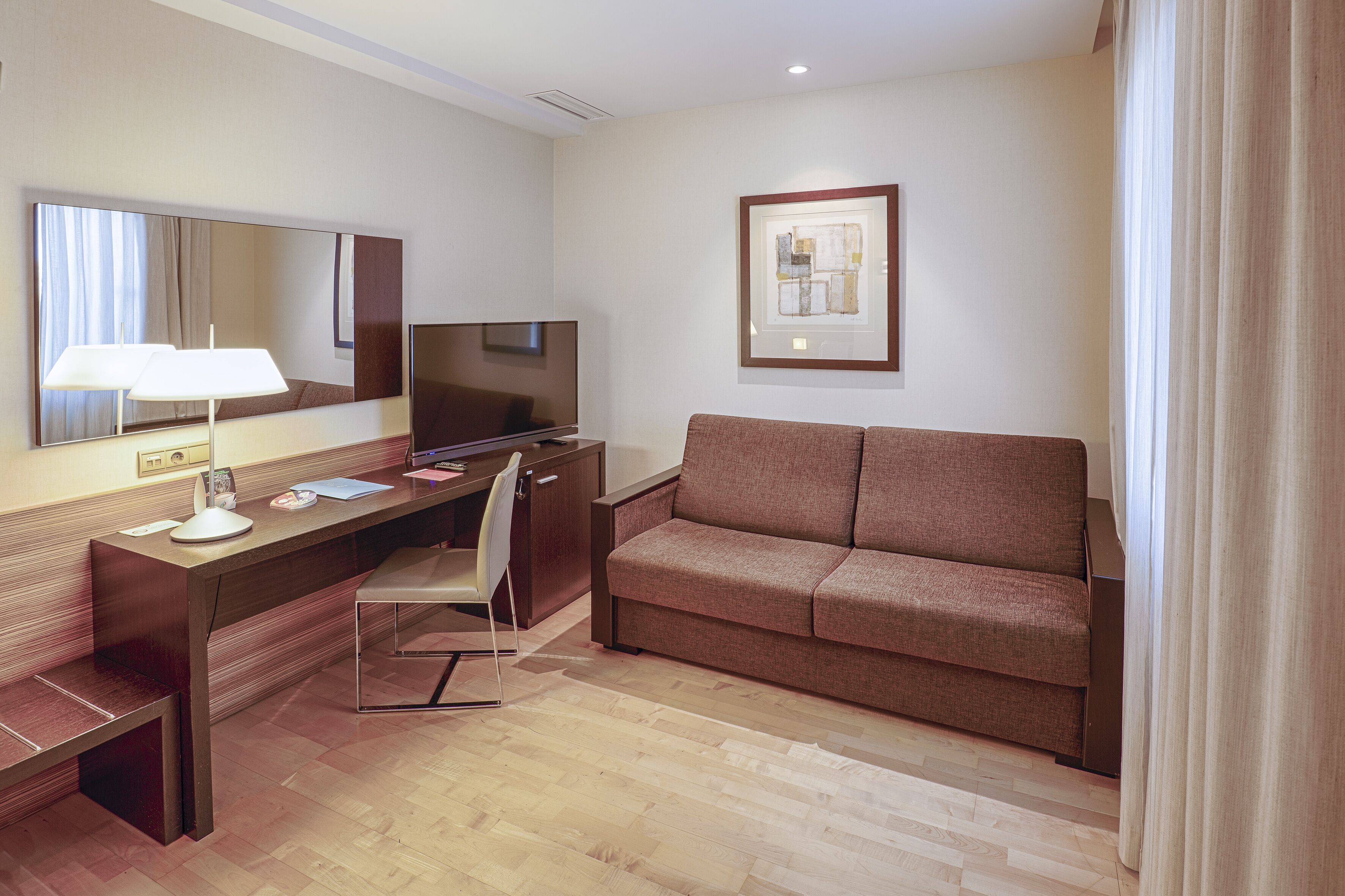 Habitación de hotel con escritorio de madera, televisión, sofá, sillón y una obra de arte enmarcada en la pared.