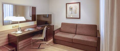 Habitación de hotel con escritorio de madera, televisión, sofá, sillón y una obra de arte enmarcada en la pared.
