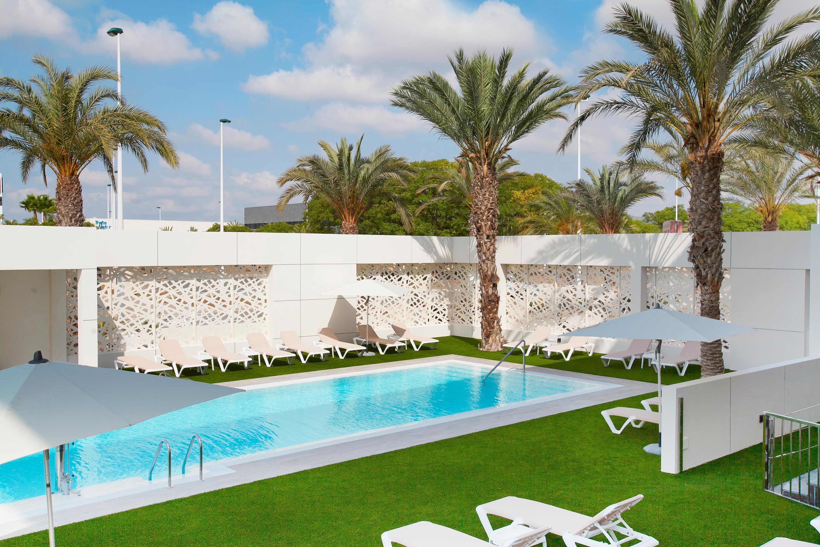 A swimming pool with white lounge chairs and umbrellas, surrounded by palm trees and a decorative white wall.