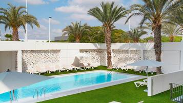 A swimming pool with white lounge chairs and umbrellas, surrounded by palm trees and a decorative white wall.
