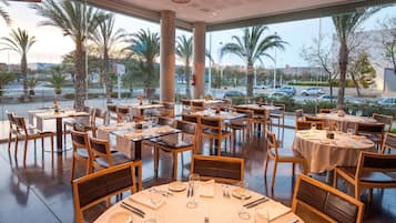 Un restaurante con varias mesas y sillas, amplios ventanales que dan a vistas de palmeras y un estacionamiento.