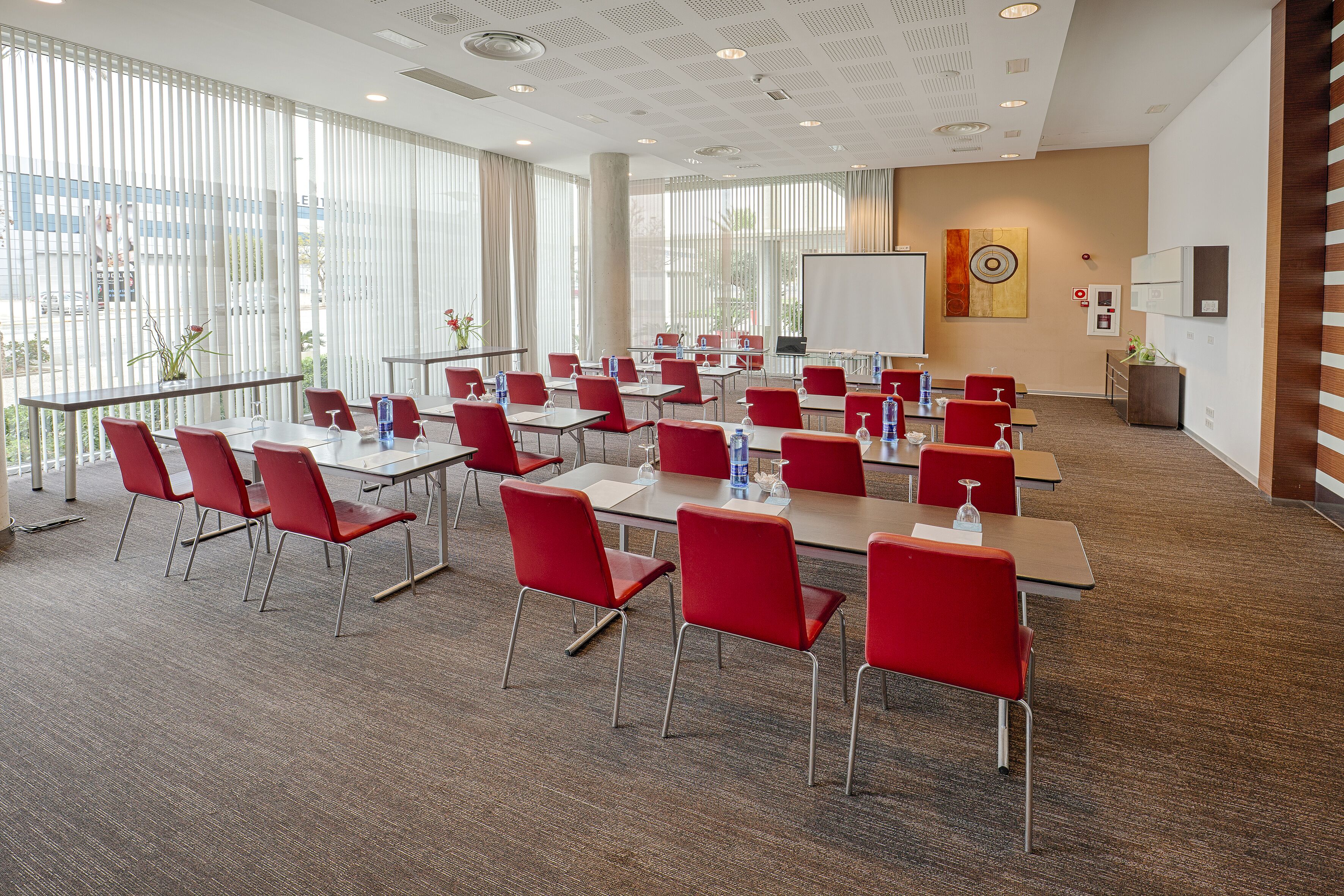 Une salle de conférence avec des chaises et des tables rouges, de grandes fenêtres et un tableau blanc.