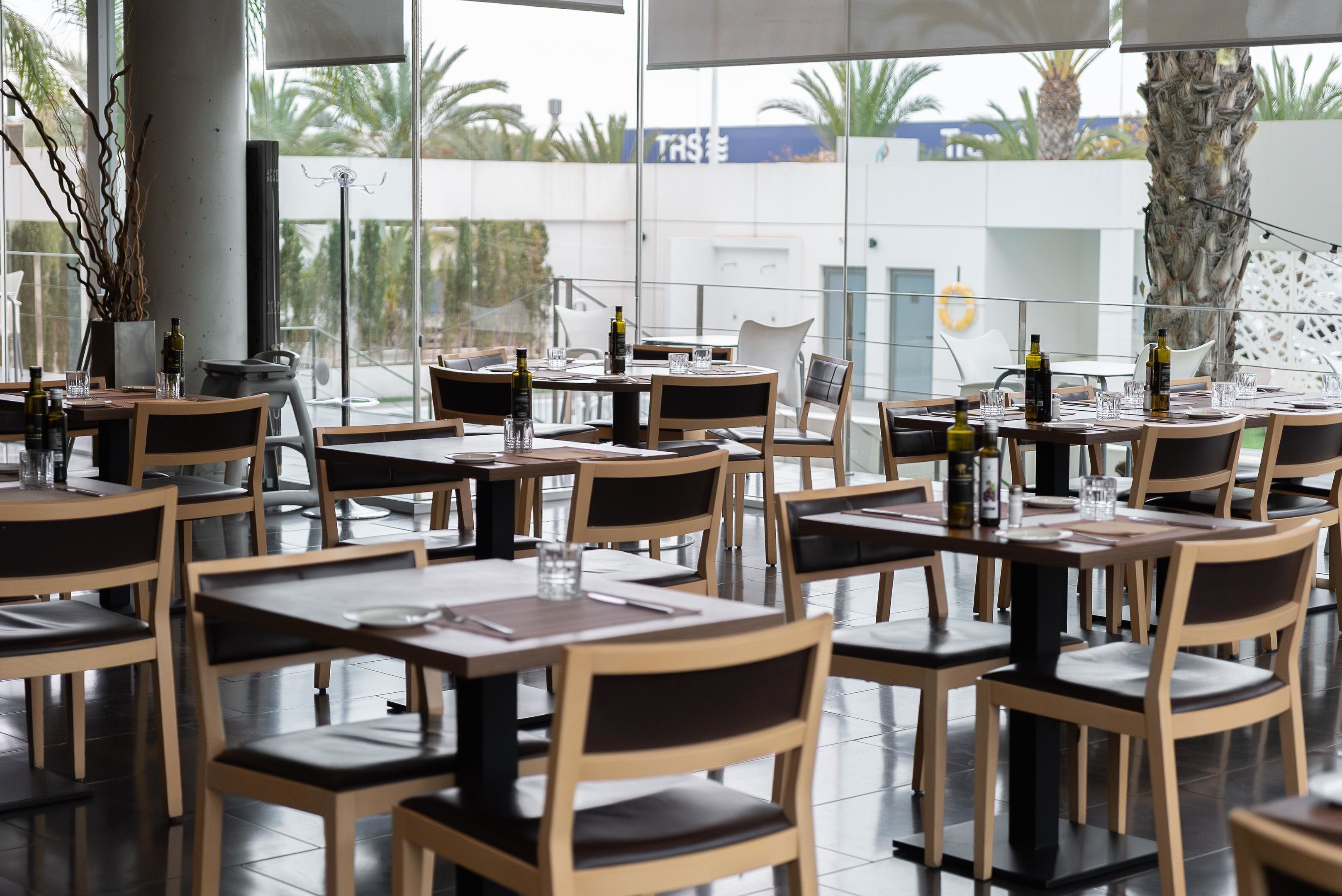 A restaurant with multiple tables and chairs, large windows, and a view of palm trees and a building with a sign.