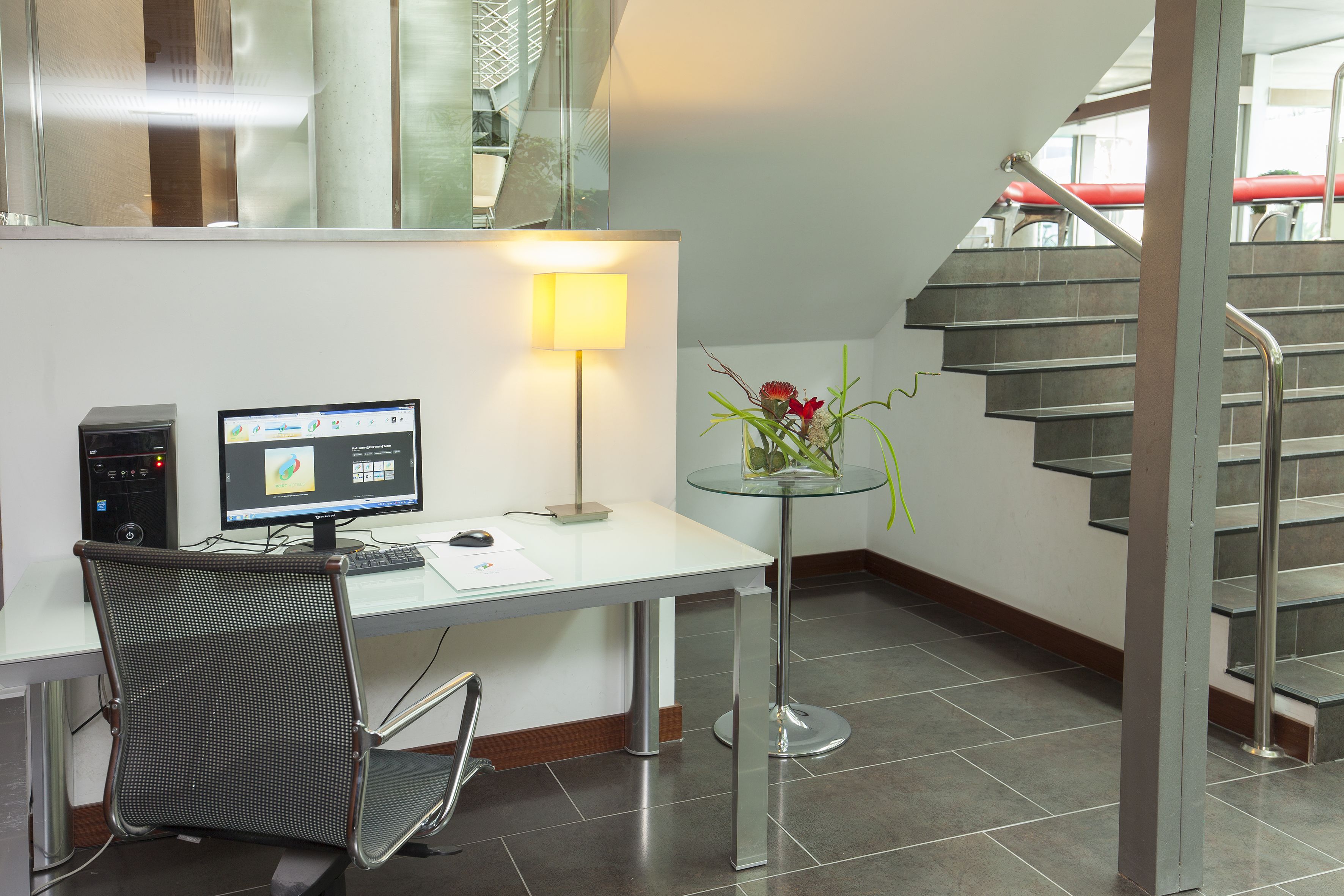 Um espaço de escritório moderno com escrivaninha, computador, cadeira, luminária e um vaso com flores.