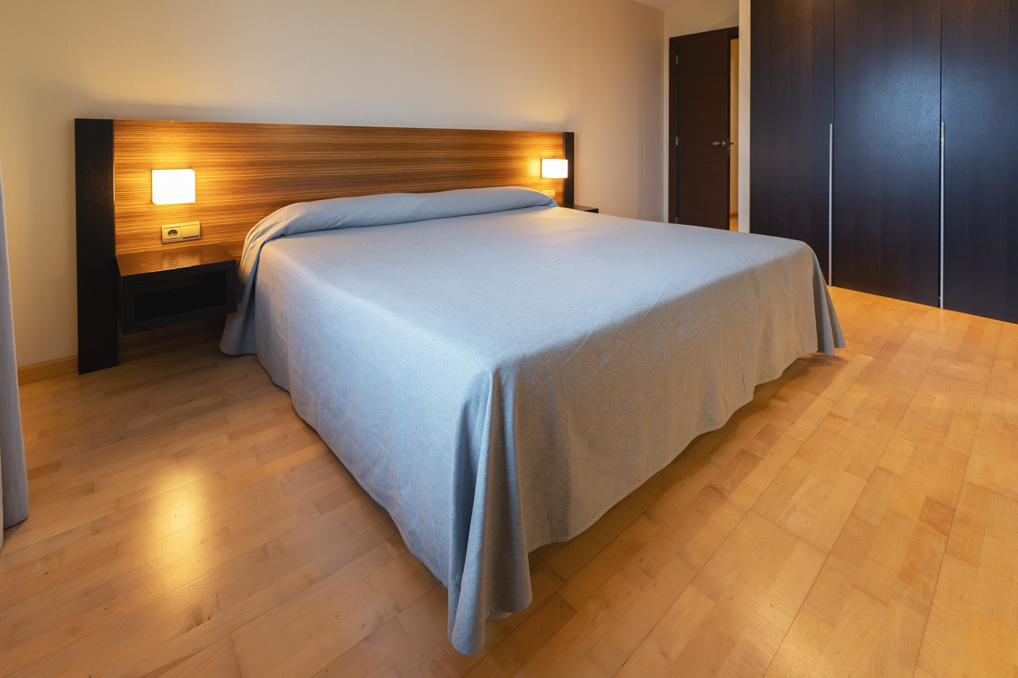 Ein ordentlich bezogenes Bett mit Holz-Kopfstück und Nachttischlampen, ein Holzboden und ein dunkles Wandschrank.