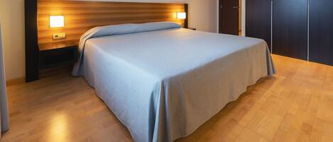 A neatly made bed with a wooden headboard and bedside lamps, a wooden floor, and a dark wardrobe.