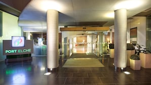 Entrada de hotel con mostrador de recepción que dice "PORT ELCHE", pilares grandes, puerta de cristal y suelo de baldosas.