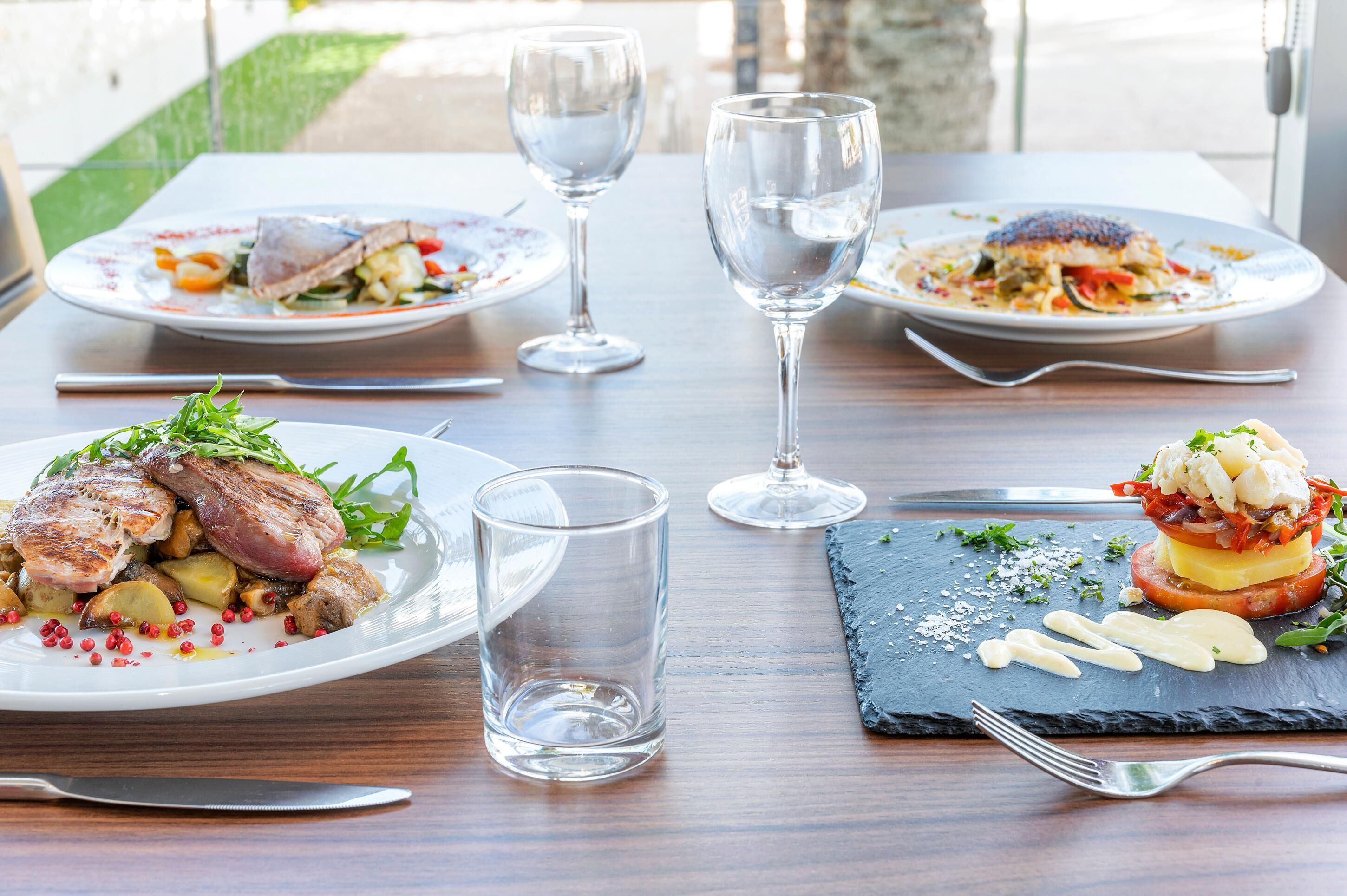 Una mesa de comedor con tres platos de comida, dos copas de vino y cubiertos.