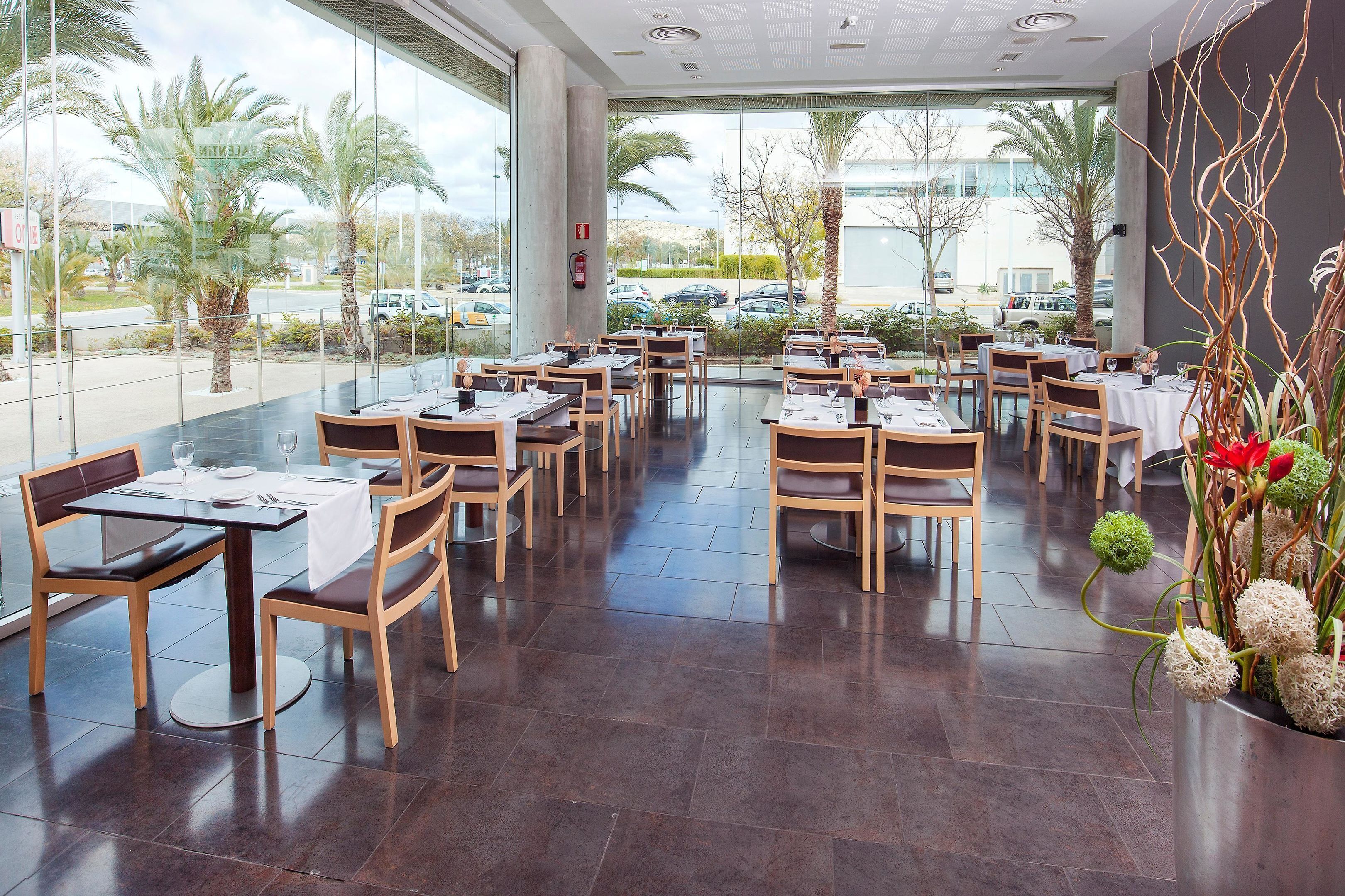 Un restaurant au décor moderne, avec des carreaux de céramique foncée au sol, des tables et des chaises en bois, et de grandes fenêtres donnant sur des palmiers et des voitures garées à l’extérieur.