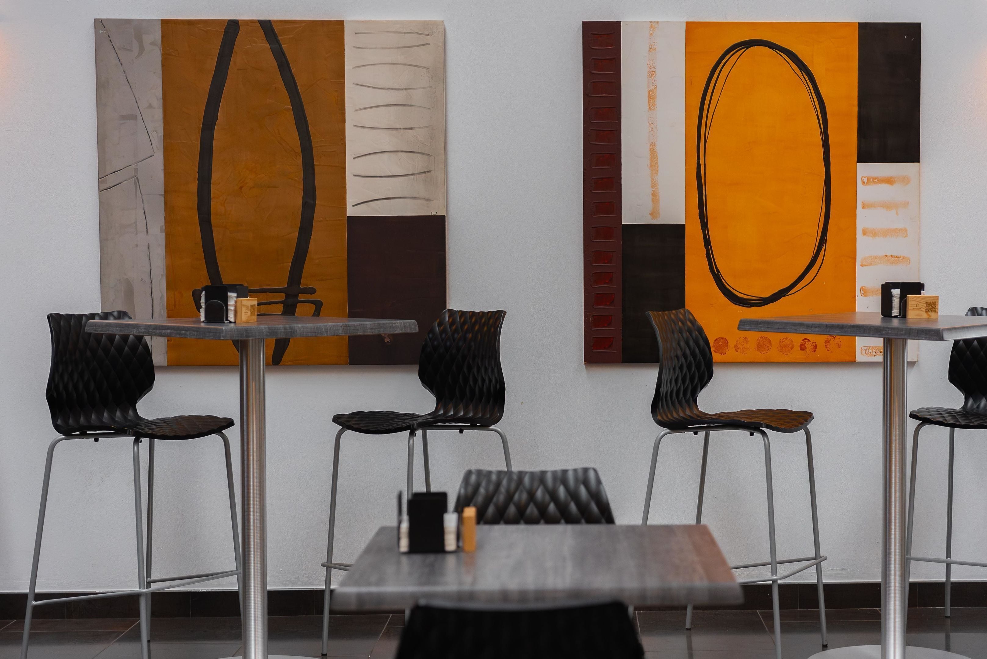 Un restaurant moderne, avec un intérieur au décor minimaliste, comprenant des chaises et des tables noires recouvertes de tissu, et des œuvres d’art abstraites accrochées aux murs.