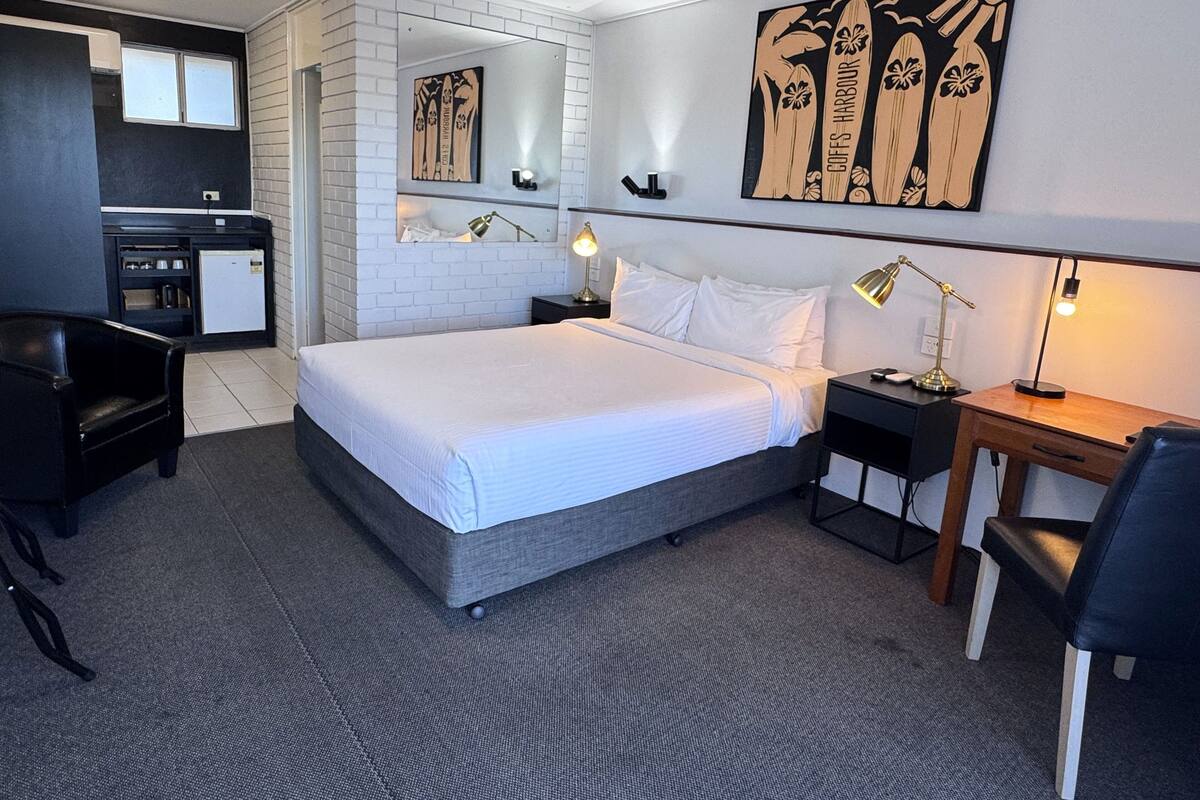A hotel room with a bed, a desk, a chair, a small kitchenette, and artwork on the walls.
