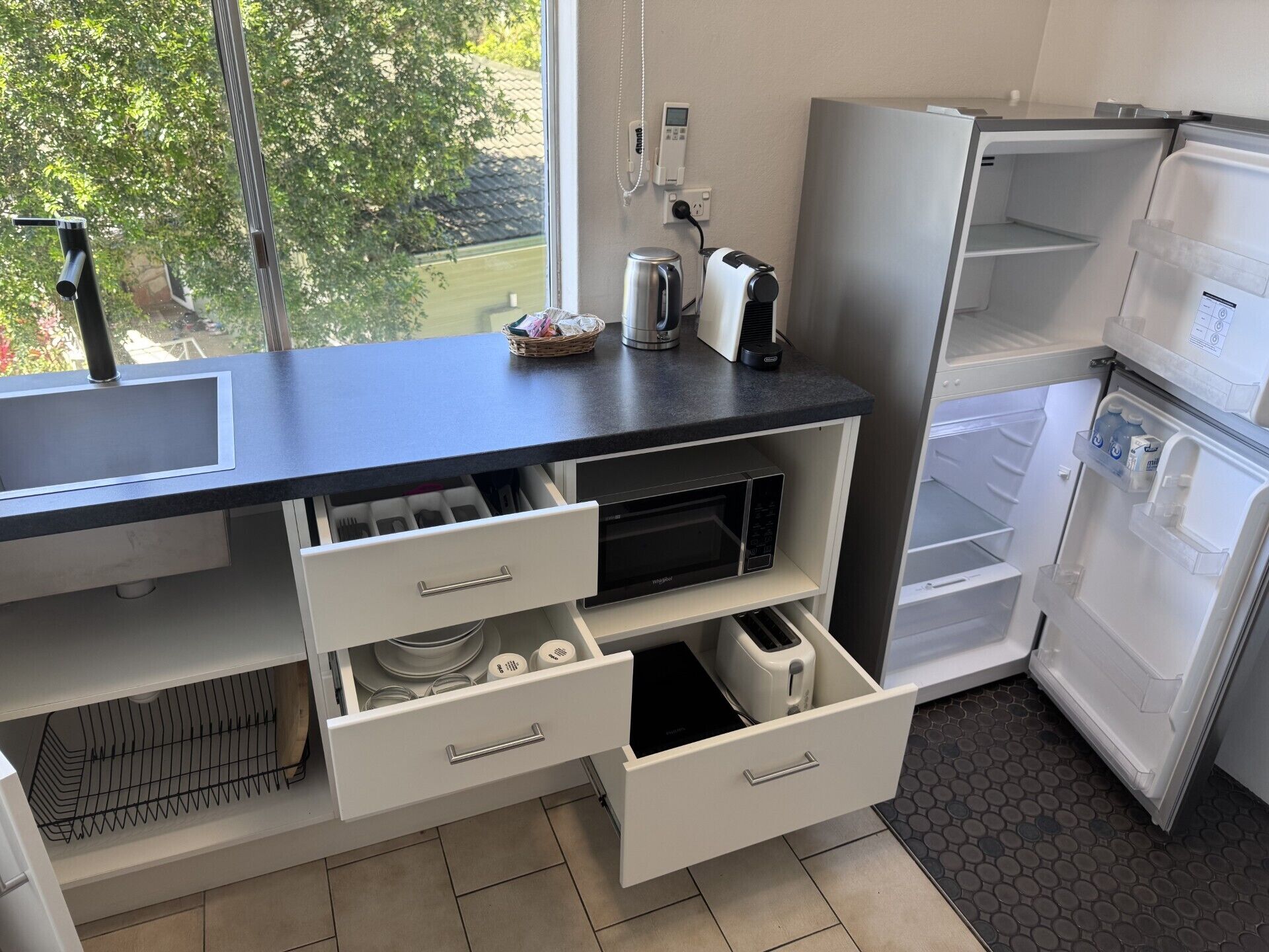 Una cocina moderna con microondas, tostadora y hervidor en la encimera, un refrigerador con dispensador de agua y una ventana con vistas a zonas verdes.
