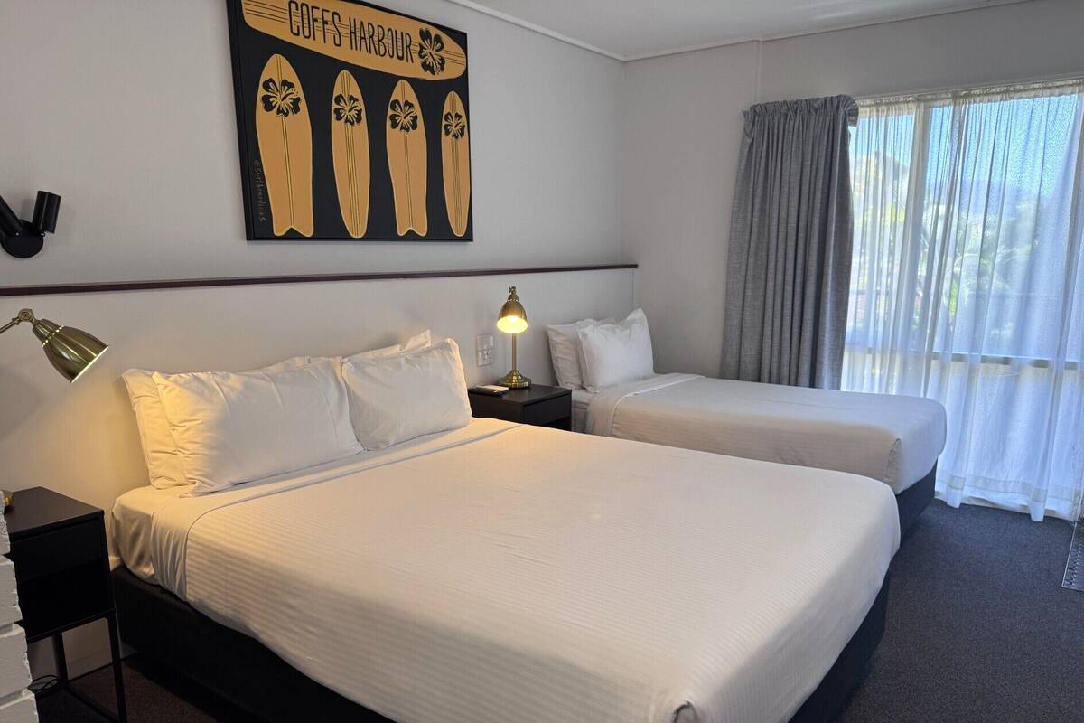 A hotel room with two beds, a large artwork of surfboards, a window with curtains, and a nightstand with a lamp.