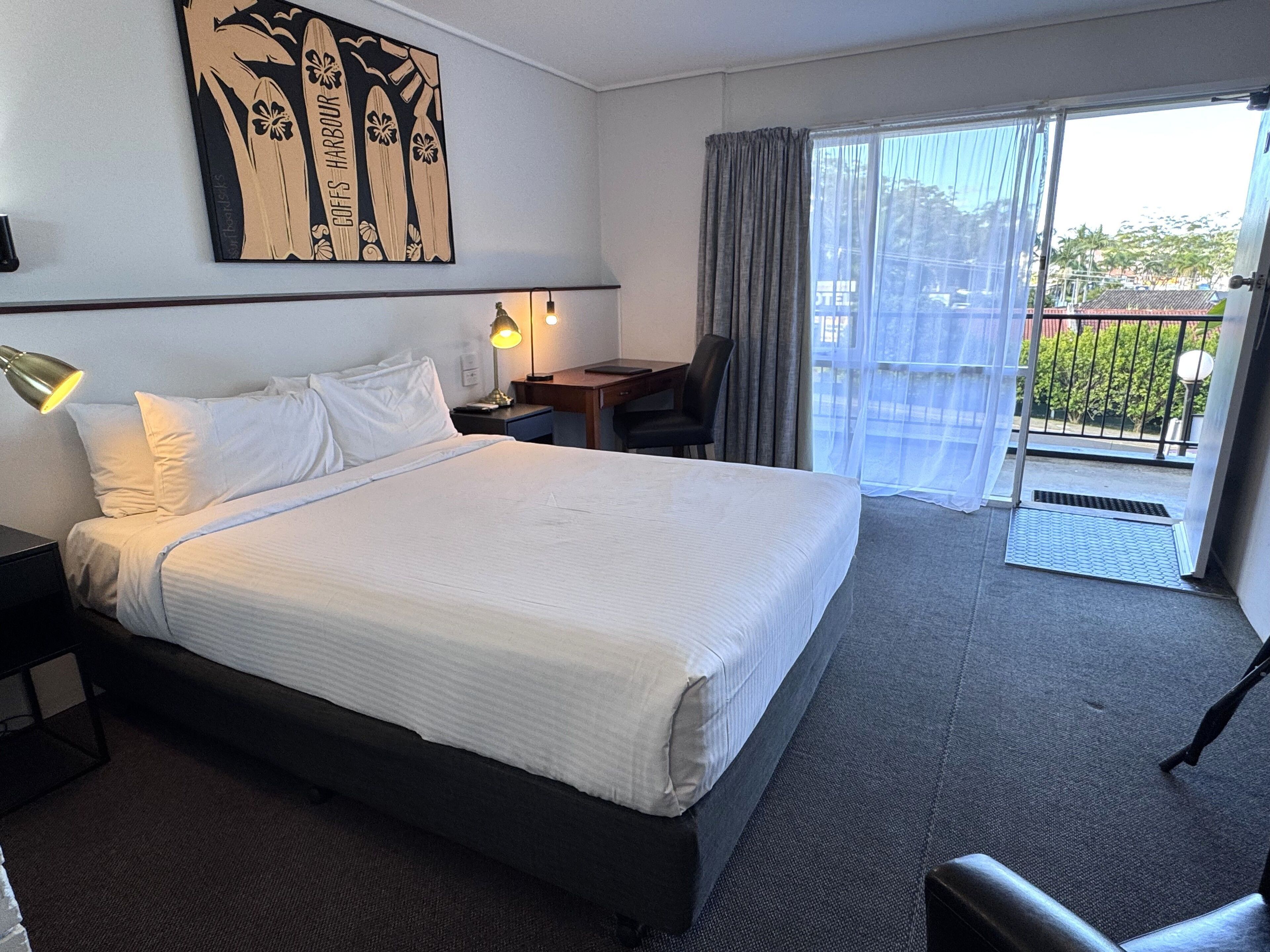 Habitación de hotel con una cama grande, un escritorio, una silla, una lámpara y vistas al exterior a través de una puerta corredera.