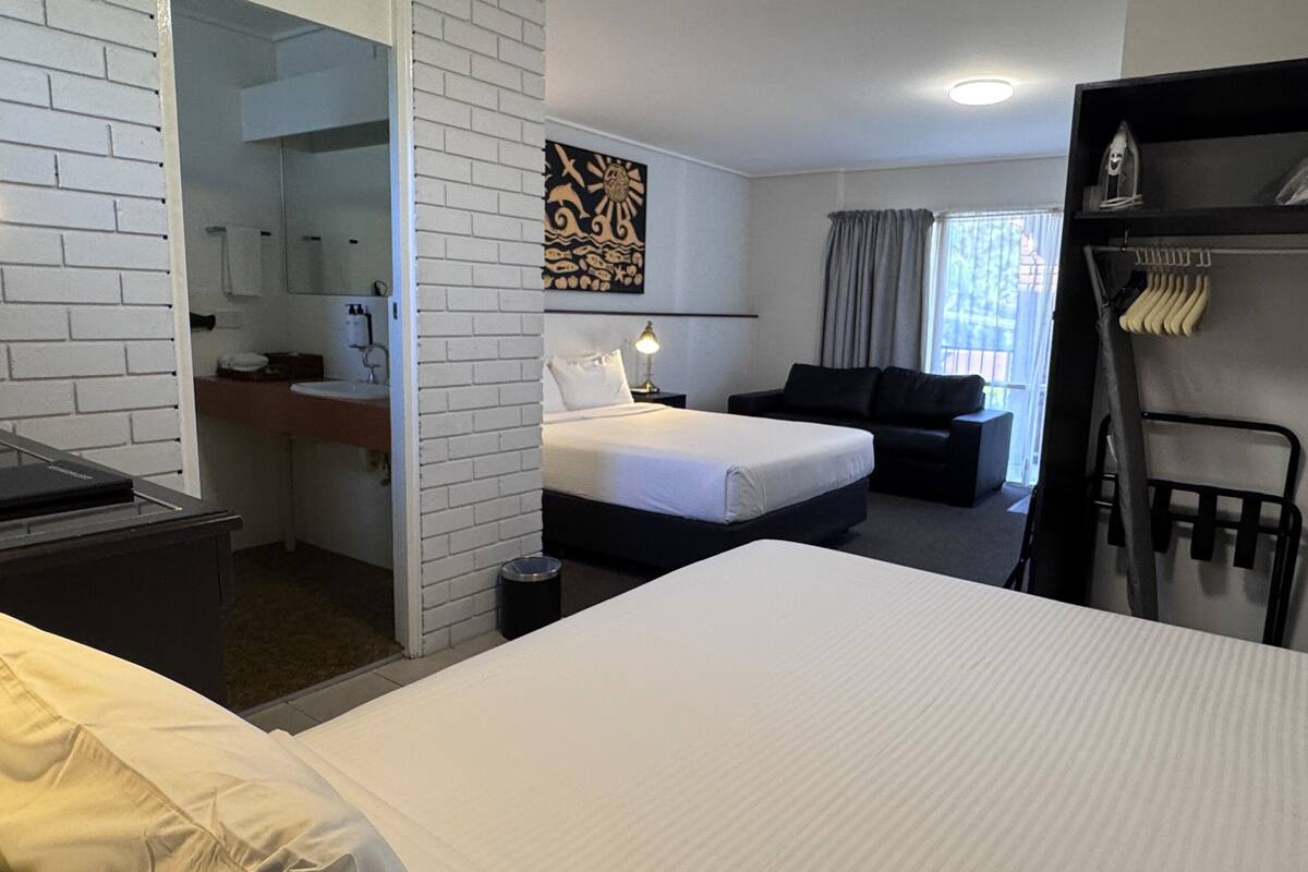 A hotel room with a bed, a desk, a chair, a black sofa, and a wall with a decorative piece.
