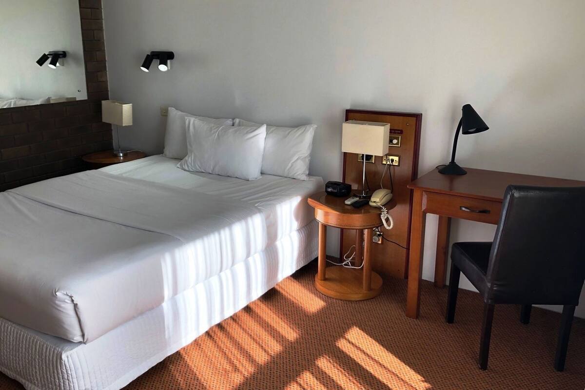 A hotel room with a bed, bedside table, desk, chair, lamp, and a wall-mounted light fixture.