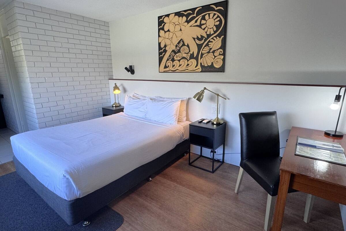 A hotel room with a bed, a desk, a chair, a nightstand, and a wall art piece.