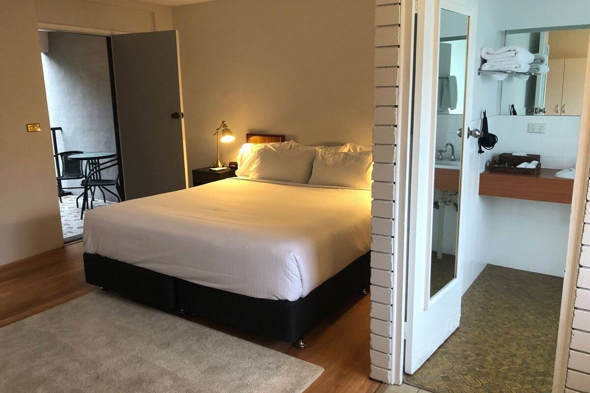 A bedroom with a bed, bedside table, lamp, and a view into the bathroom.