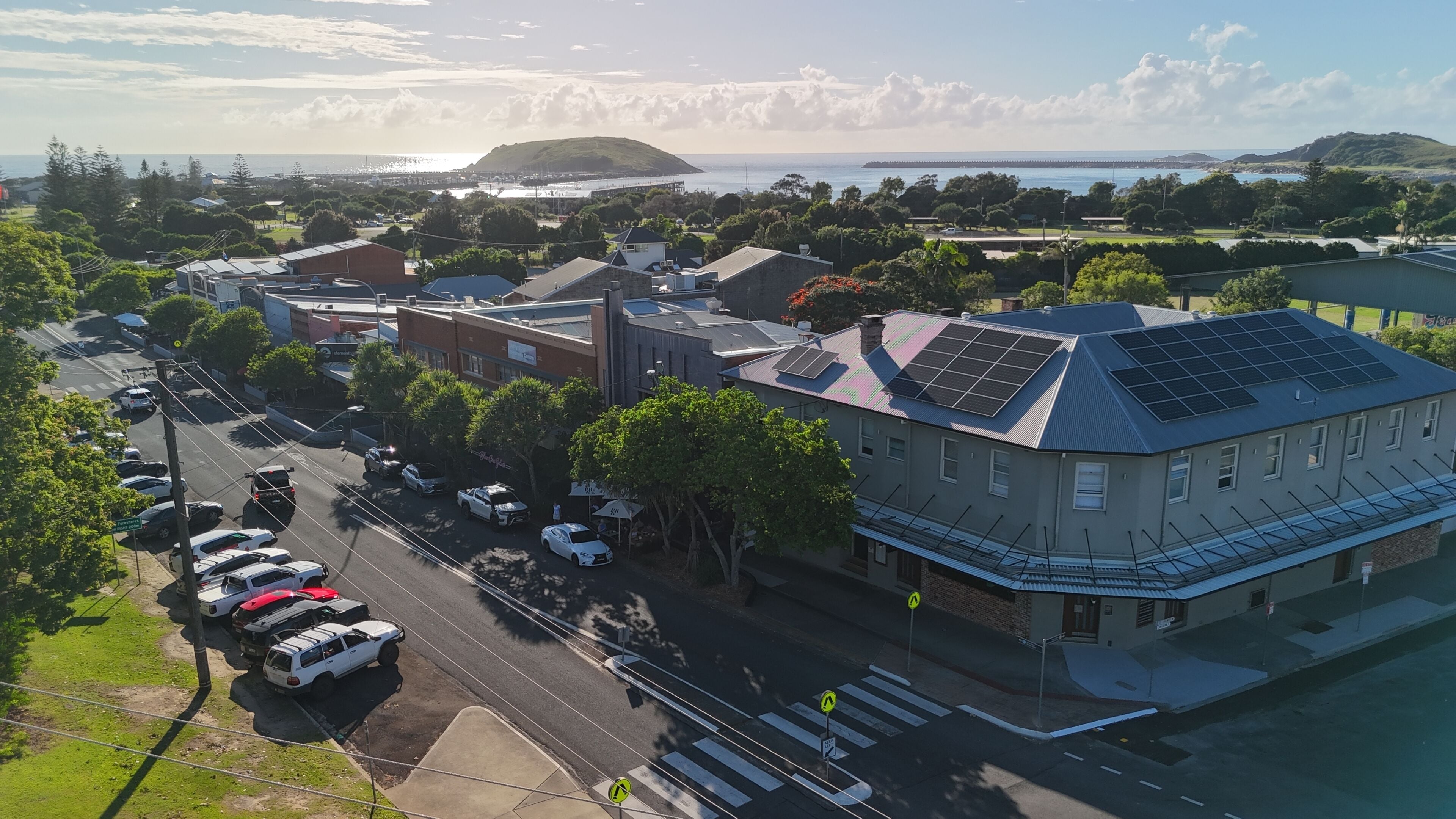 A coastal town with a prominent building featuring solar panels, a street with parked cars, and a clear view of the sea.