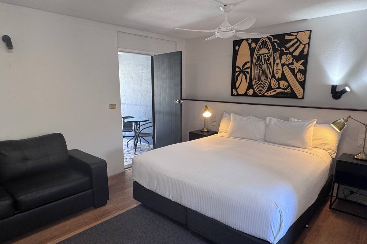 A bedroom with a bed, a black sofa, a ceiling fan, a wall art piece, and a small table with a lamp.