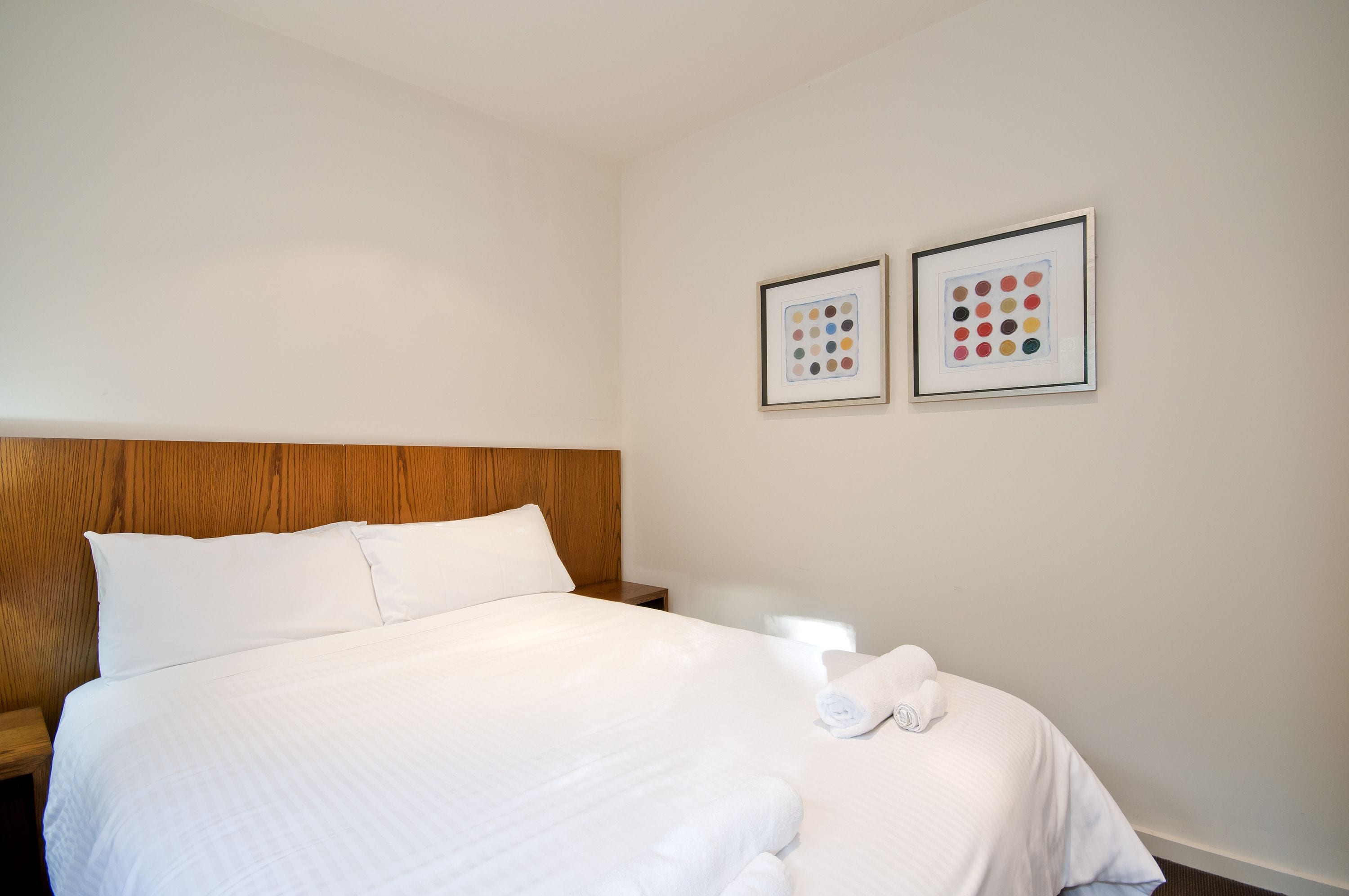 Un lit bien fait, avec du linge de lit blanc, une tête de lit en bois et deux œuvres d’art encadrées accrochées au mur.