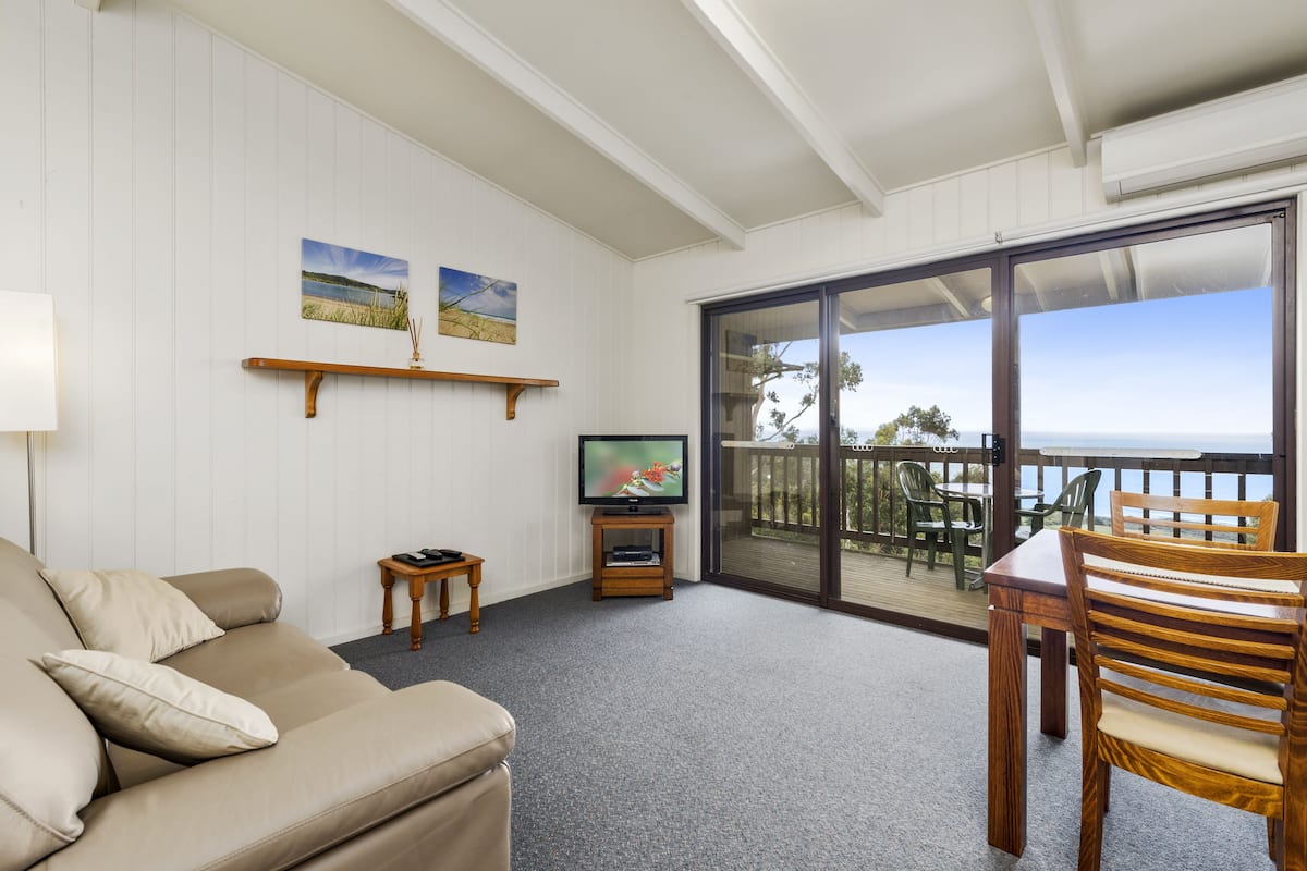 Un salon avec un canapé beige, un téléviseur posé sur un meuble en bois et un balcon offrant une vue sur la mer.