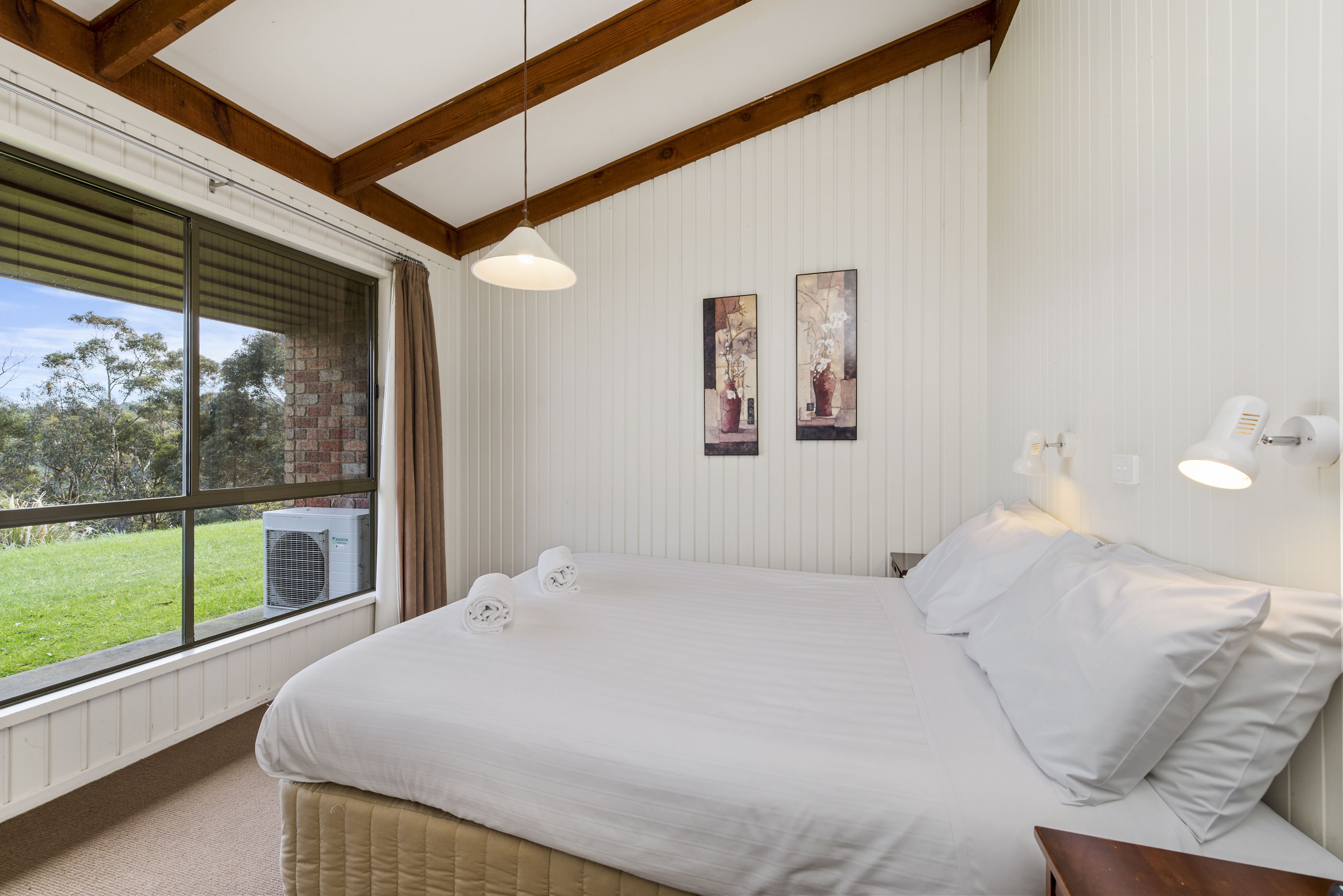 A bedroom with a large bed, a window with blinds, a wall-mounted air conditioner, and two framed pictures.