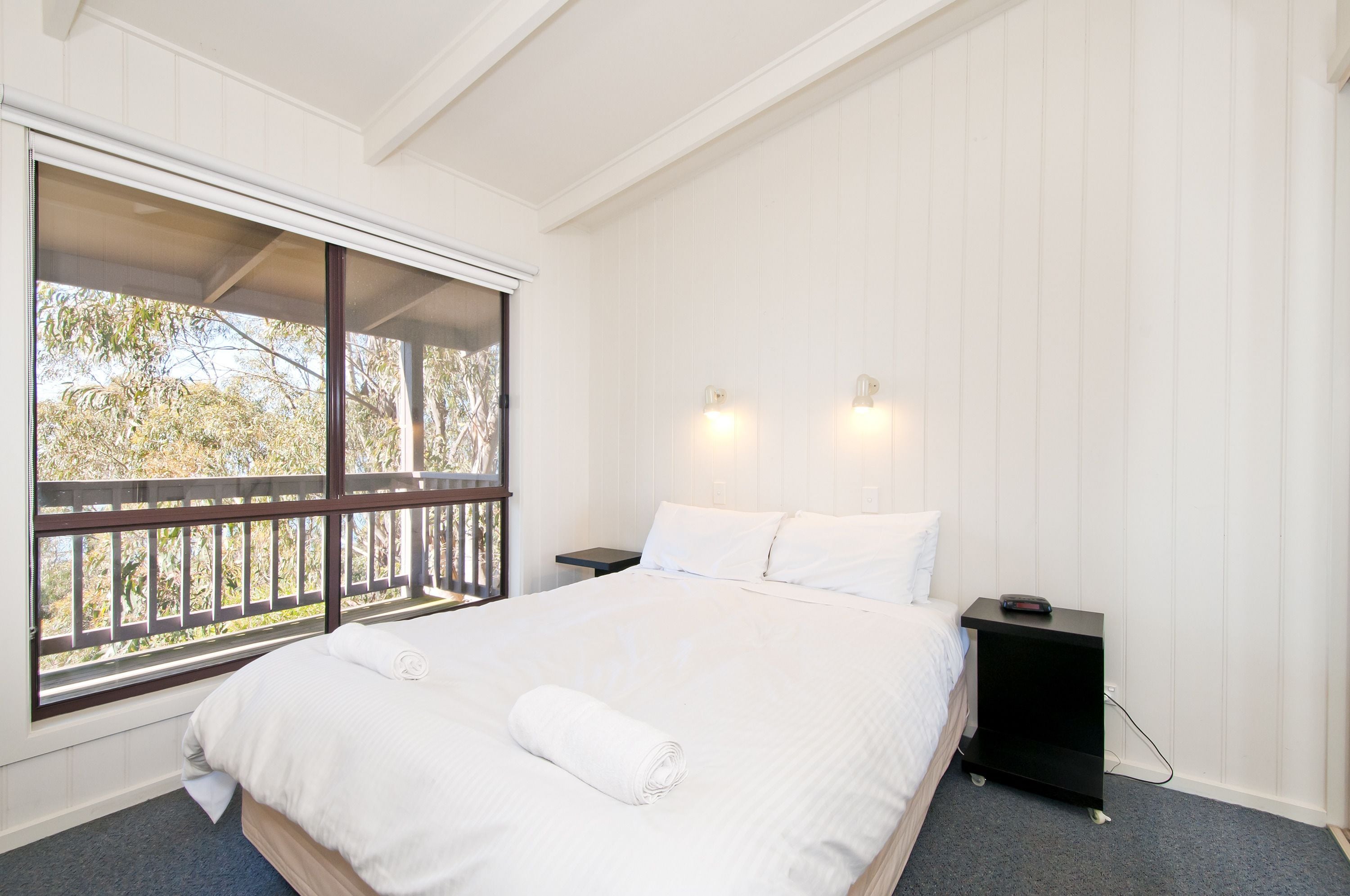 A bedroom with a large window, a bed with white linens, two bedside lamps, and a small black nightstand.