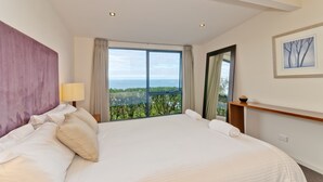 A large bed with a view of the sea, a nightstand with a lamp, and a framed picture on the wall. - Beacon Point Ocean View Villas (Skenes Creek)