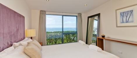 A large bed with a view of the sea, a nightstand with a lamp, and a framed picture on the wall.