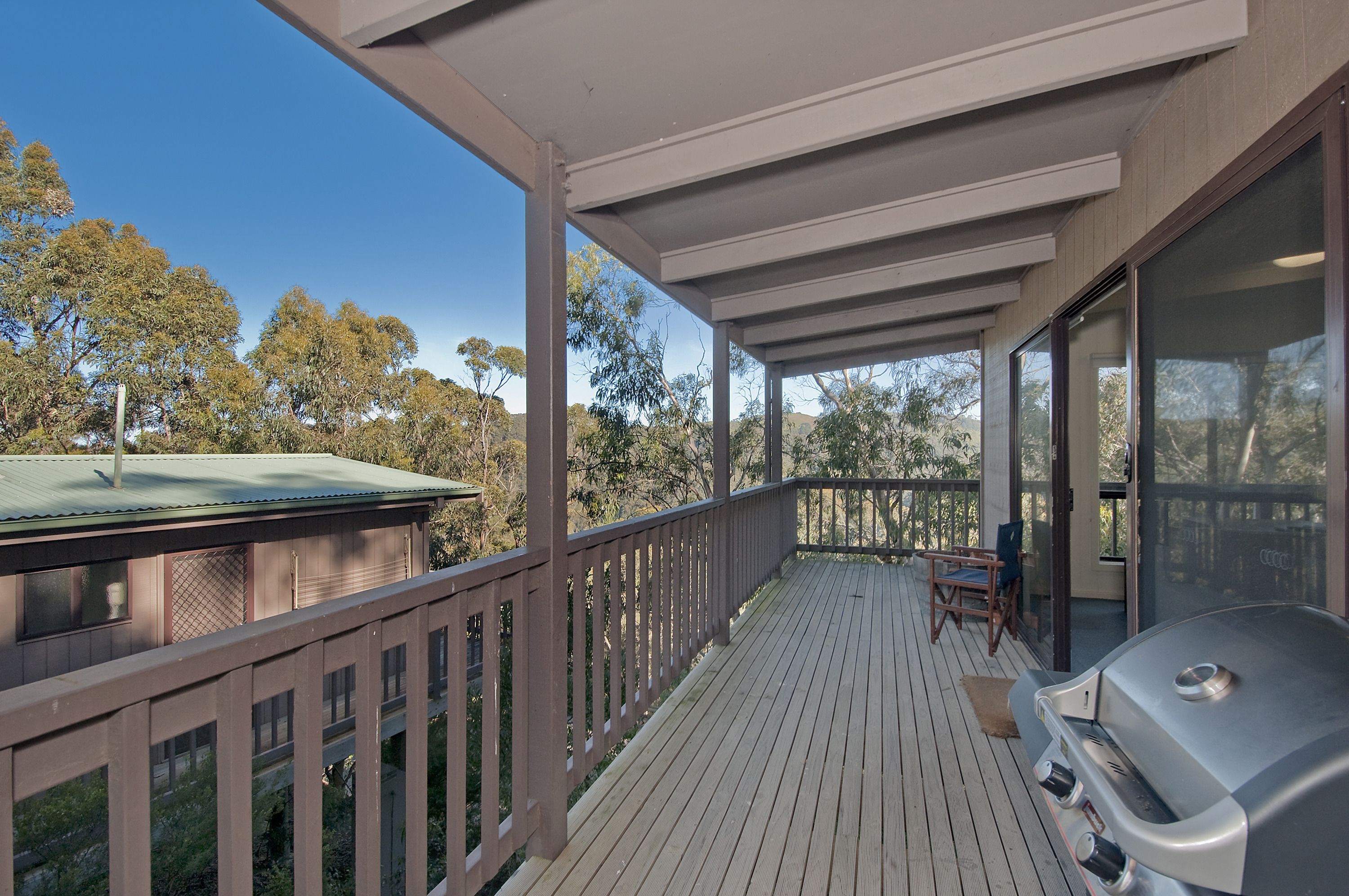 A covered outdoor deck with a barbecue grill, wooden railings, and a view of trees.