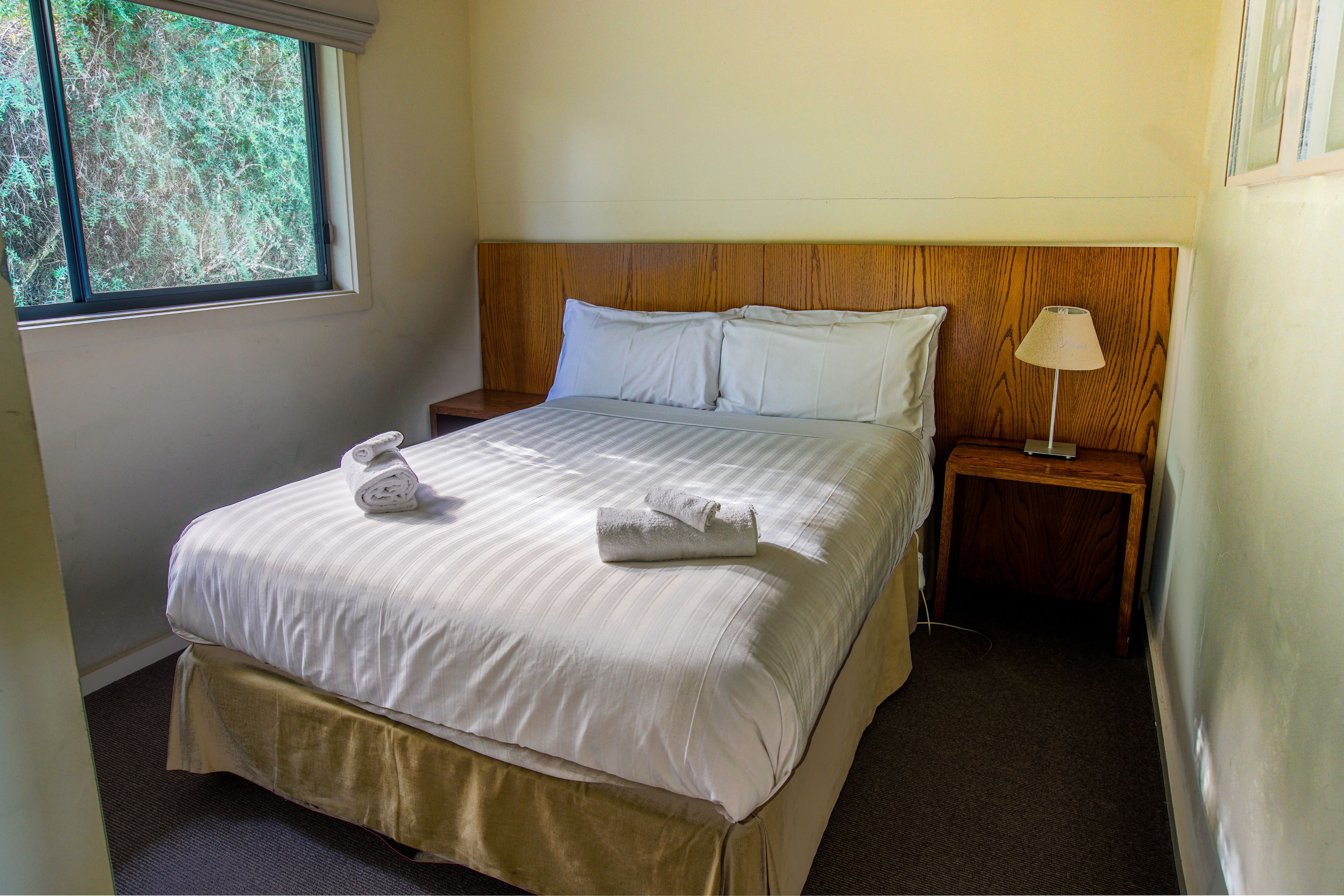 A neatly made bed with white linens and two rolled towels, a wooden headboard, a bedside table with a lamp, and a window with a view of trees.