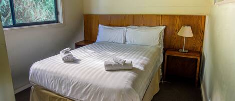 A neatly made bed with white linens and two rolled towels, a wooden headboard, a bedside table with a lamp, and a window with a view of trees.