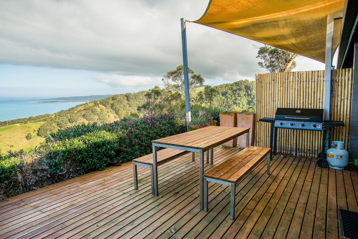 Une terrasse en bois avec une table et un banc, un barbecue et une vue sur l’océan et les collines.