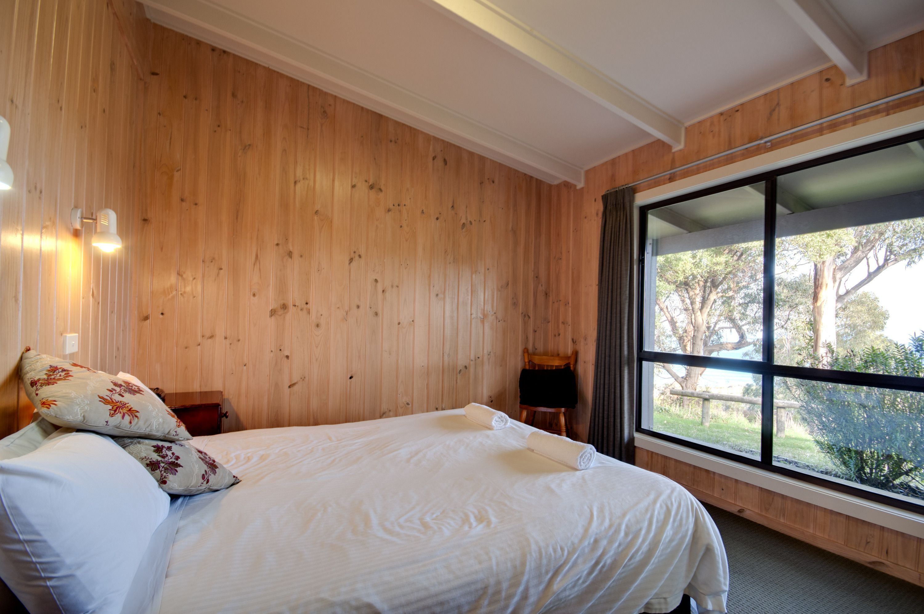 A bedroom with a wooden wall, a large window, and a bed with white bedding.