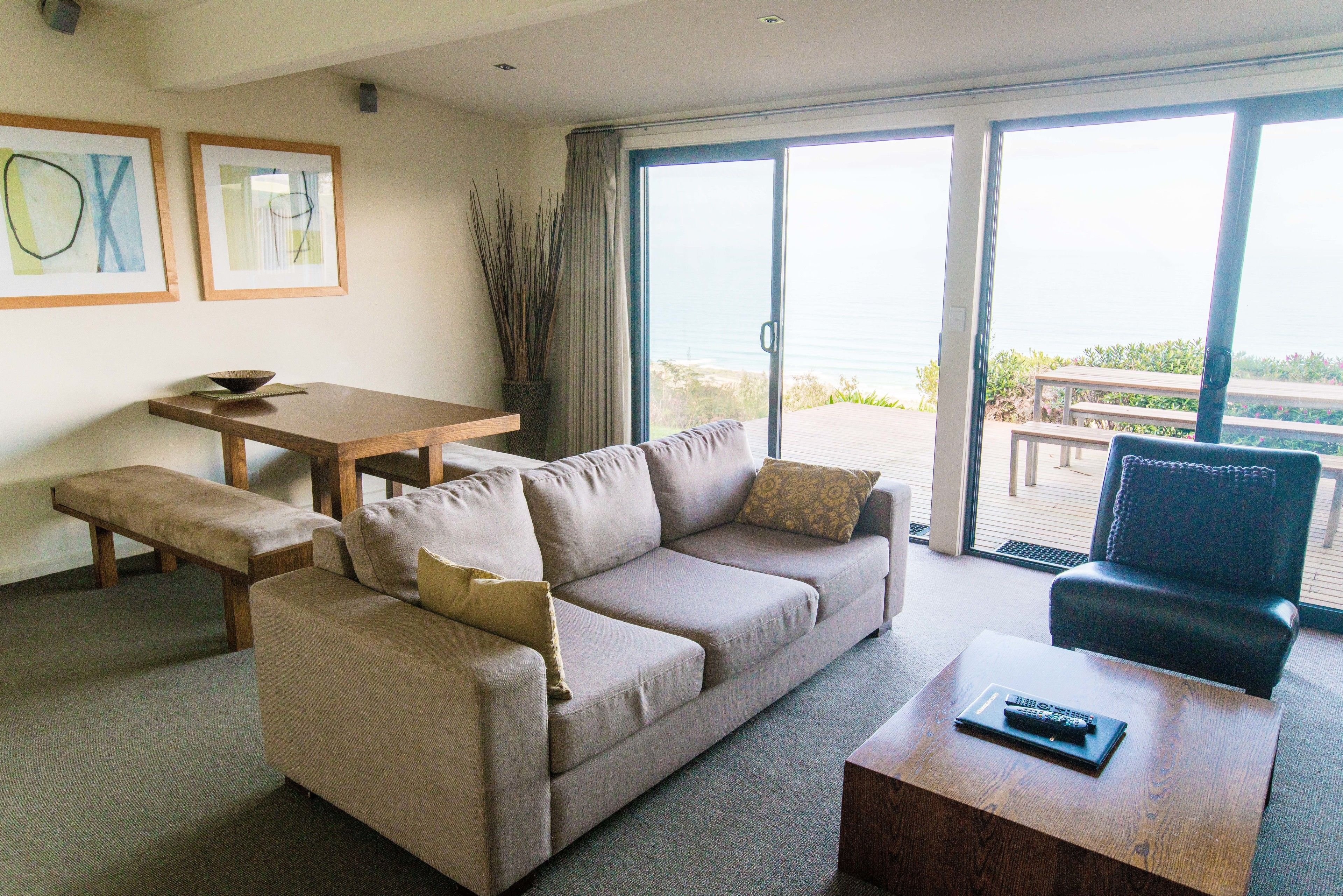 A living room with a sofa, a coffee table, a bench, a dining table, and a view of the outdoors through large sliding doors.