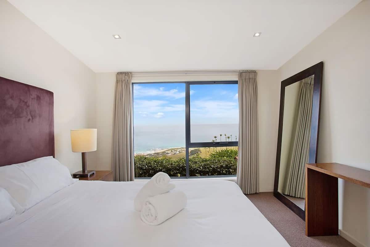 Une chambre d’hôtel avec un grand lit, une table de chevet avec une lampe, un bureau et une grande fenêtre donnant sur la plage.