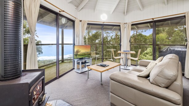 A cozy living room with a sofa, a TV, and a table. There are large windows offering a view of the outdoors. - Beacon Point Ocean View Villas (Skenes Creek)