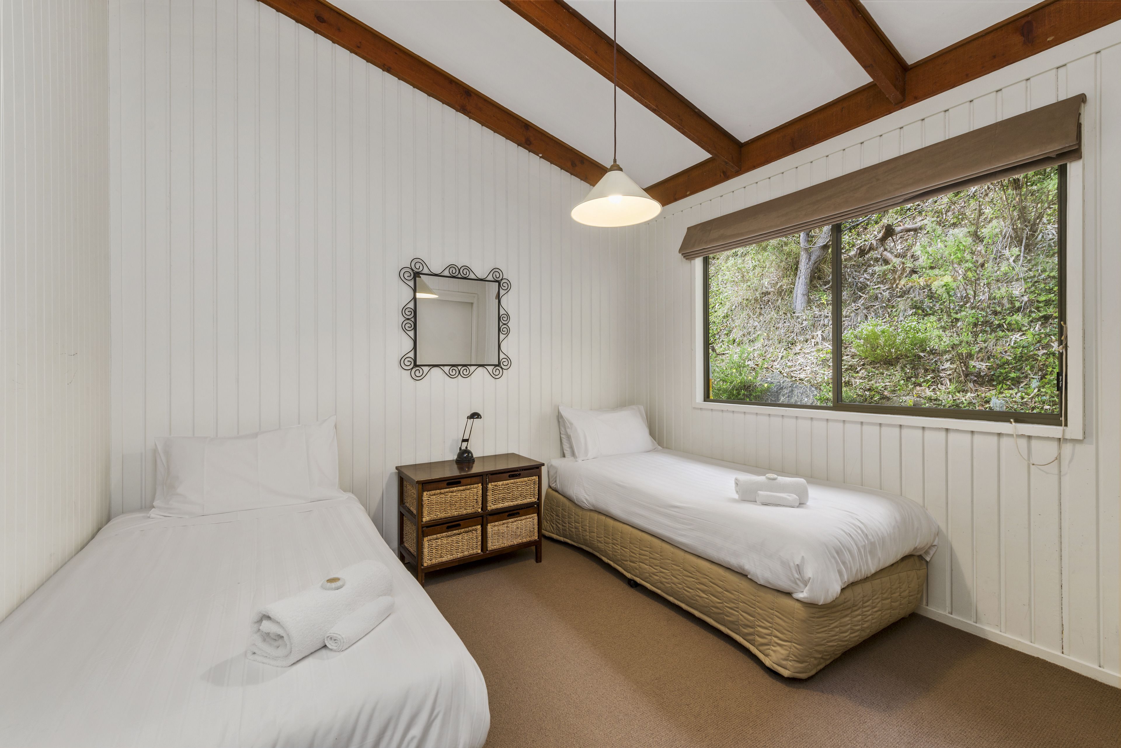 A bedroom with two beds, a nightstand, a mirror, and a window with a view of greenery.